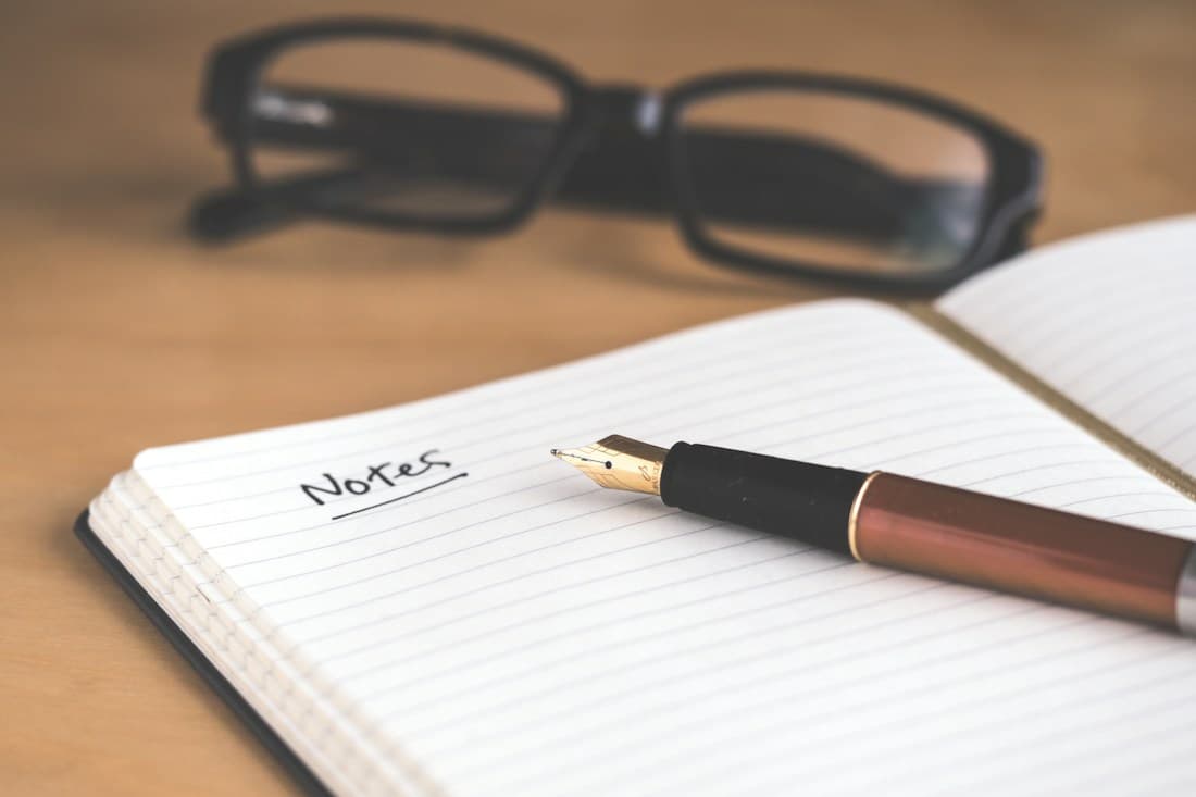 Cuaderno con la palabra Notes, una pluma y unos lentes sobre una mesa de madera