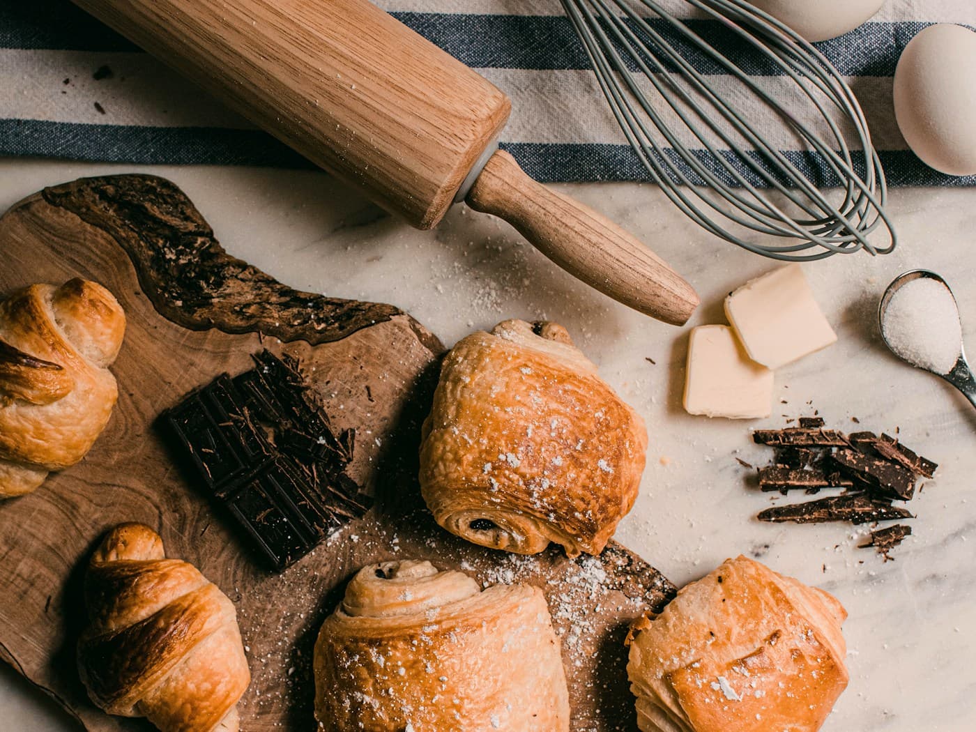 Mesa de repostería con harina, mantequilla, chocolate, croissants y un rodillo de madera