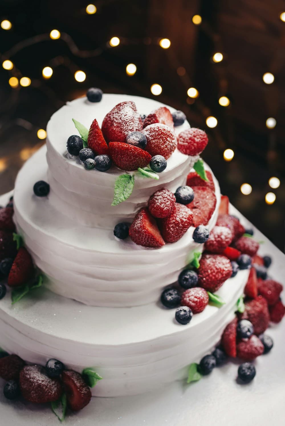 Torta blanca de tres pisos con frutillas, frambuesas y arándanos, con luces al fondo